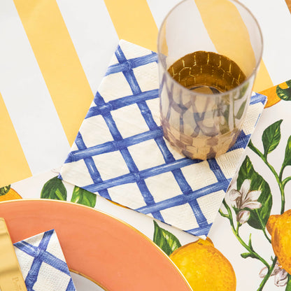 A table setting with Blue Lattice Napkins by Hester &amp; Cook in a lattice pattern, featuring China Blue ink designs, and a glass of lemon juice.
