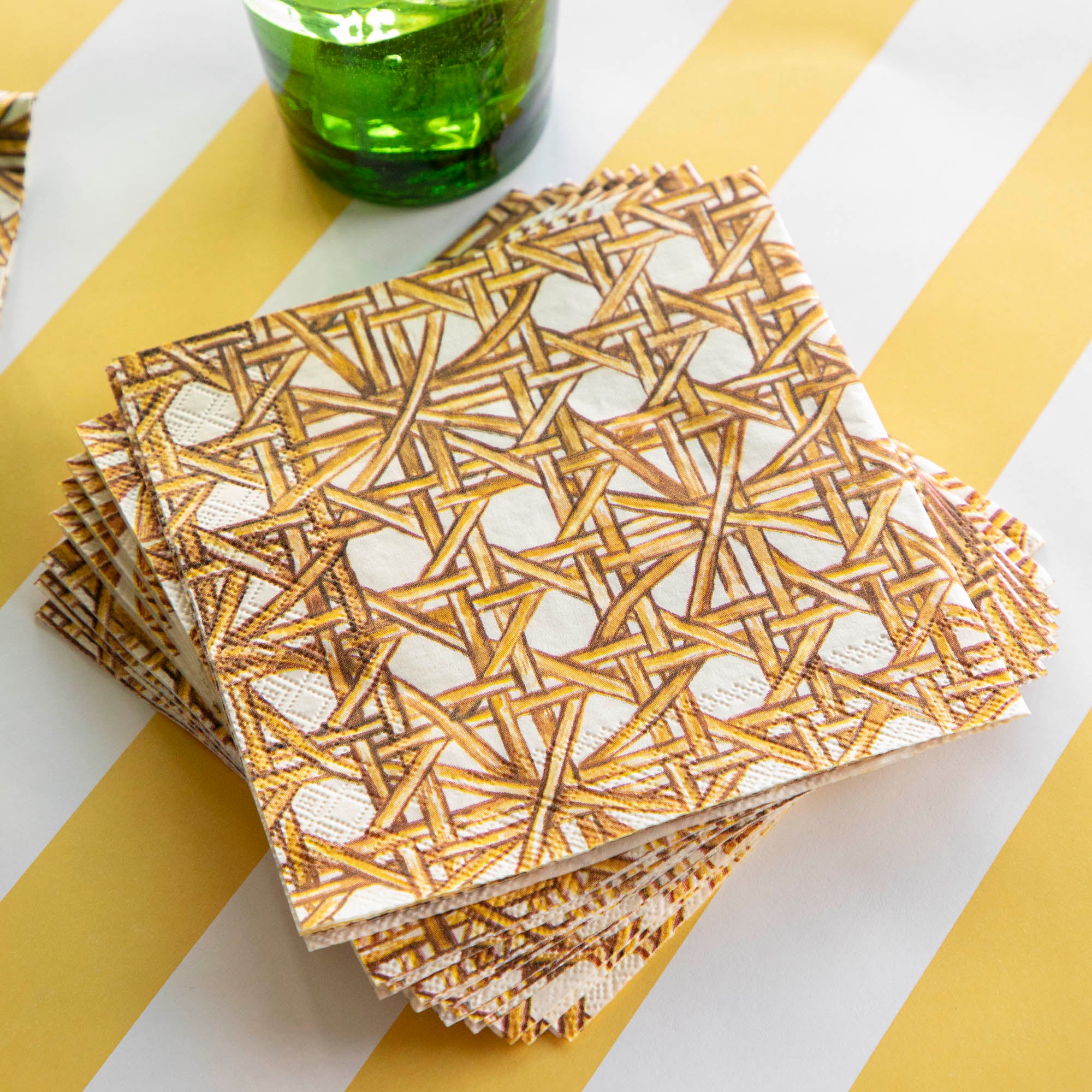 A set of Rattan Weave Napkins from Hester &amp; Cook and a bottle of beer on a yellow and white striped tablecloth, creating a visually appealing tablescape.