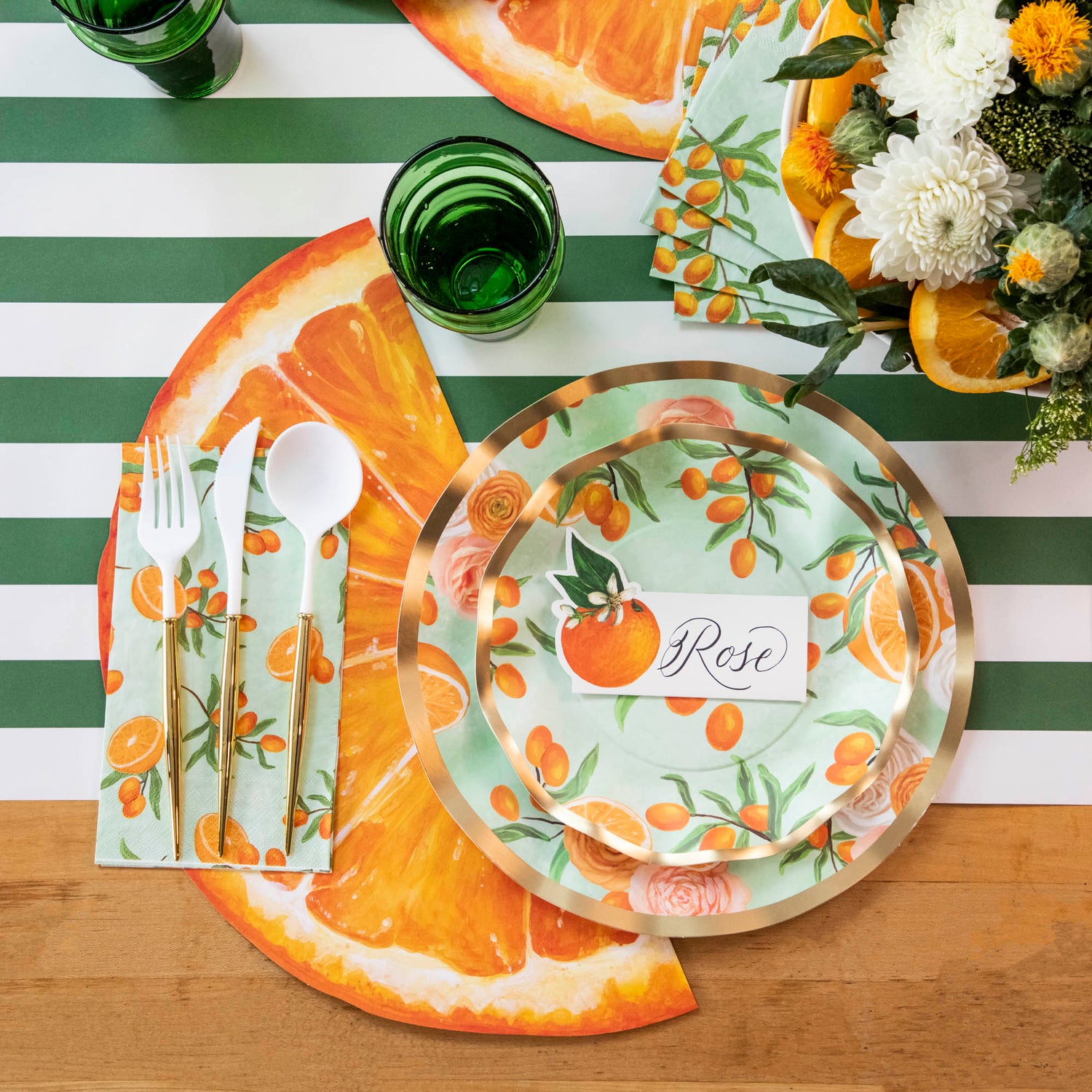 A table setting with disposable Sophistiplate White/Gold Cutlery Set and orange slices on a green and white striped tablecloth.