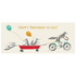 A whimsical illustration of four raccoons zooming by: one is pedaling a teal bicycle, with two holding balloons in a red wagon pulled behind the bike, and the last one hanging on to the wagon, over a cream background, with "Happy Birthday To You!" written in gold across the top of the card.