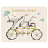 A whimsical illustration of three white and black storks riding on a green tandem bicycle over a beige background, with "CONGRATULATIONS!" printed in gold across the top of the card.