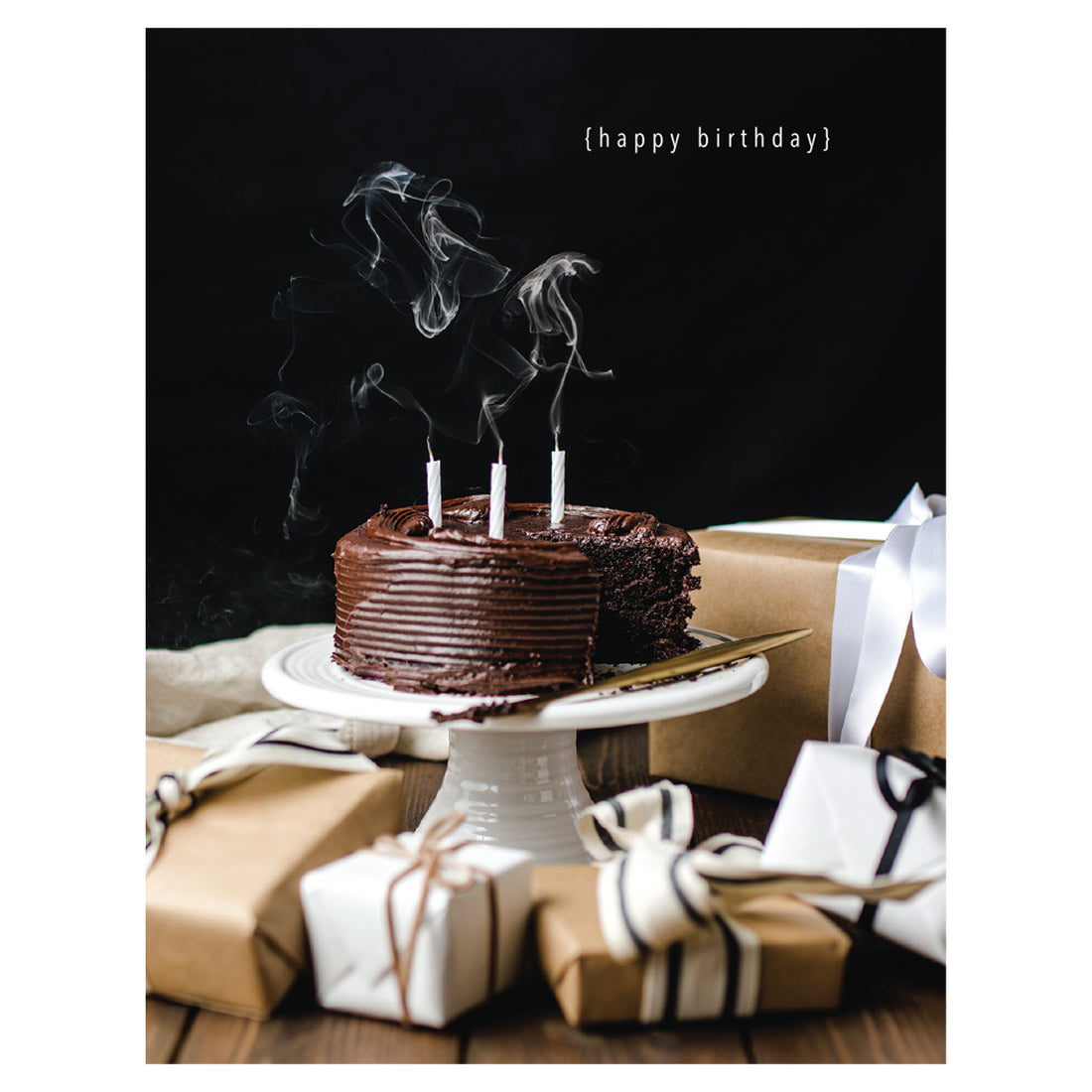A photo of a chocolate cake on a cake stand, candles freshly blown out and smokey, on a table  surrounded by kraft-wrapped gifts in white ribbons on a black background, with &quot;{happy birthday}&quot; printed in white in the upper right of the card.