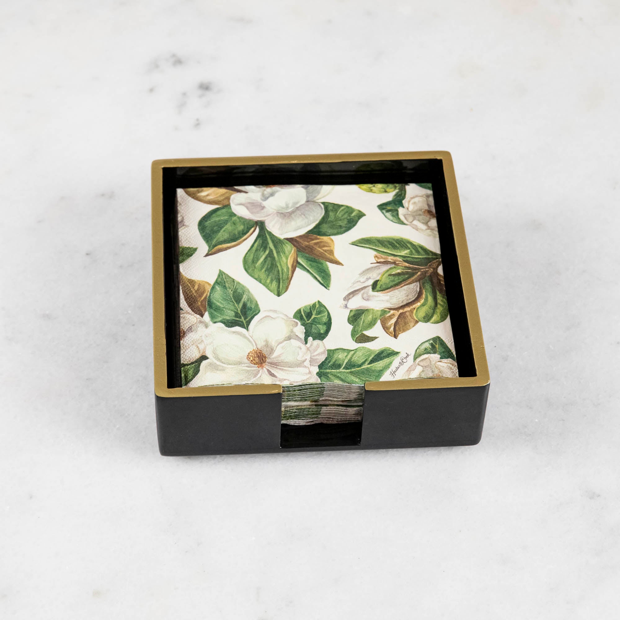 A black lacquered napkin holder with gold trim and a magnolia print napkin inside of it. 