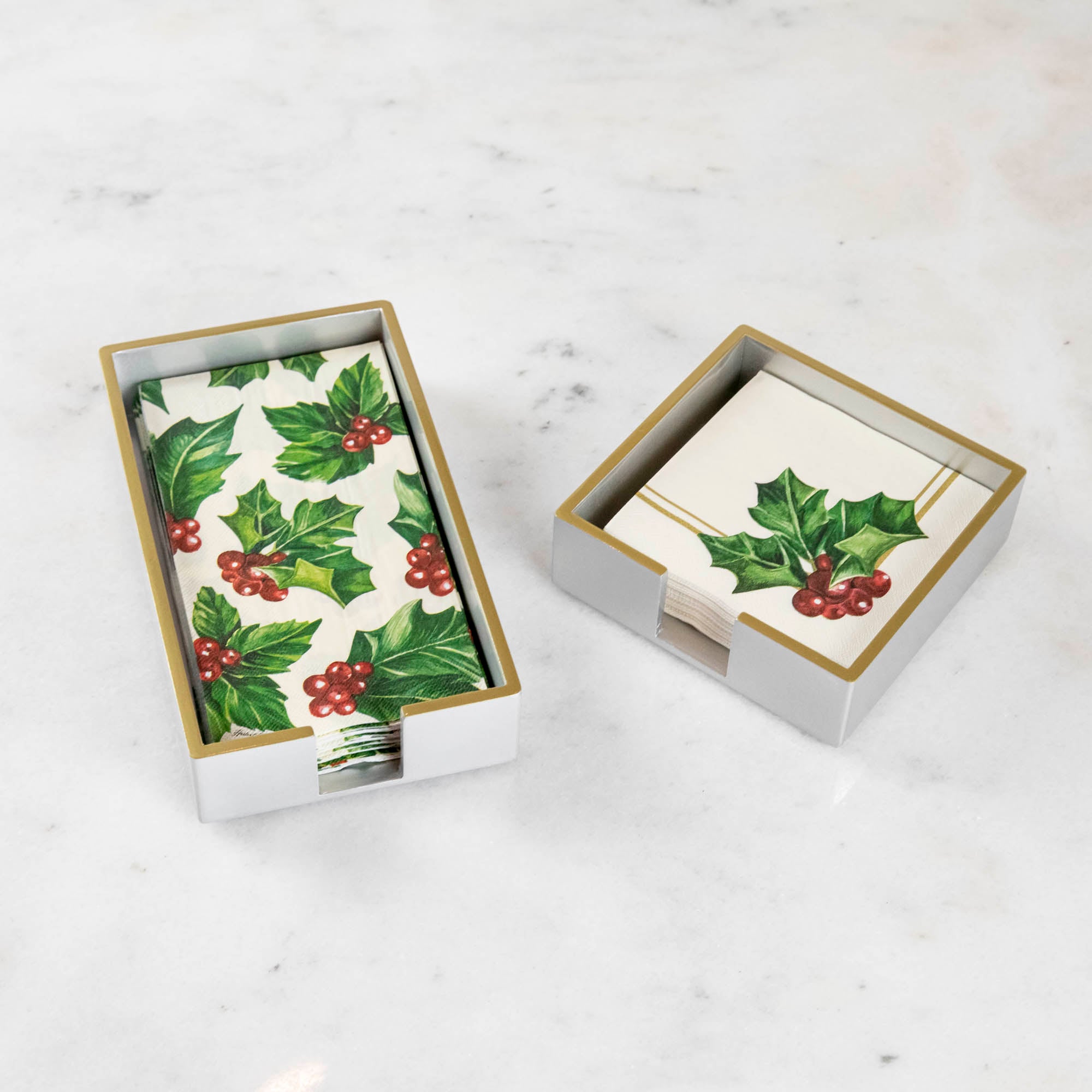 A silver colored lacquered napkin holder in guest and cocktail size with napkins in them. 