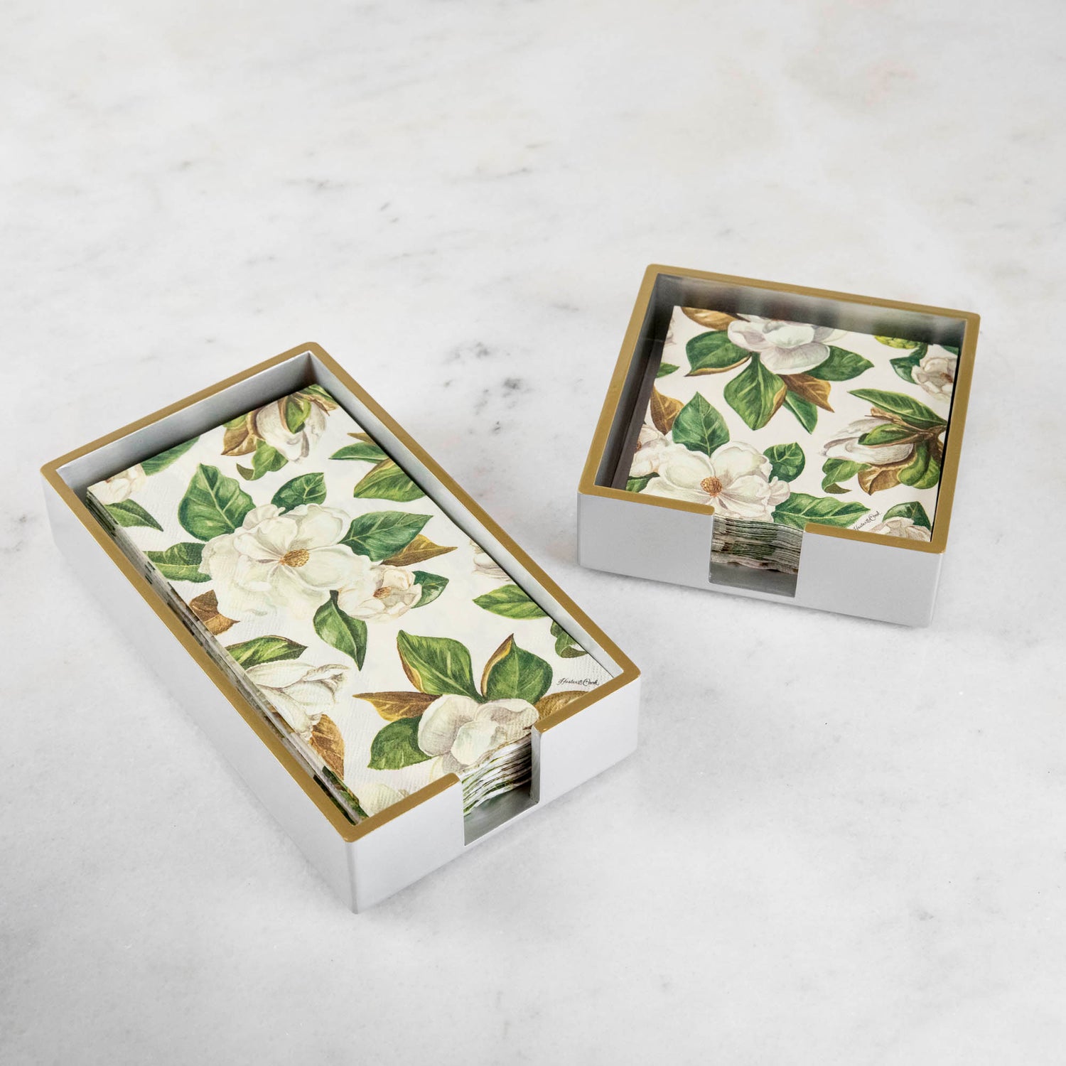 A silver colored lacquered napkin holder in guest and cocktail size with napkins in them. 