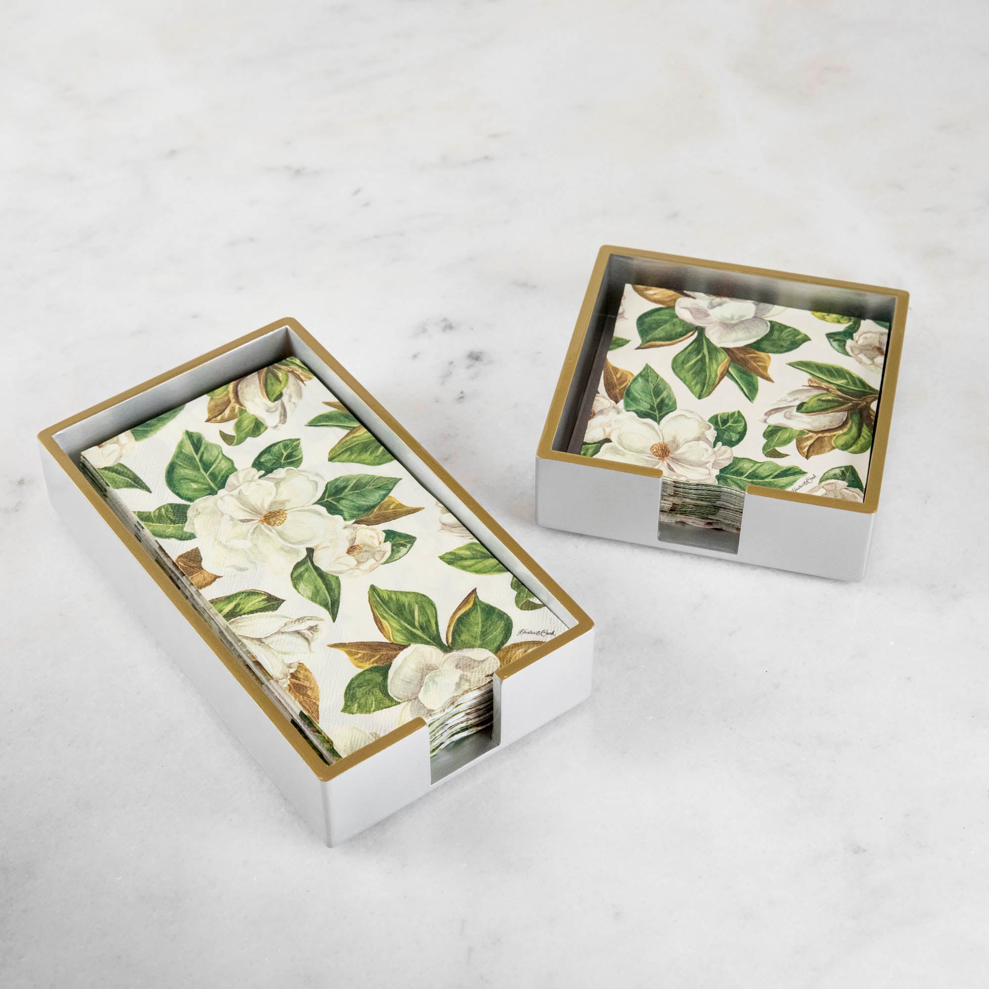 A silver colored lacquered napkin holder in guest and cocktail size with napkins in them. 