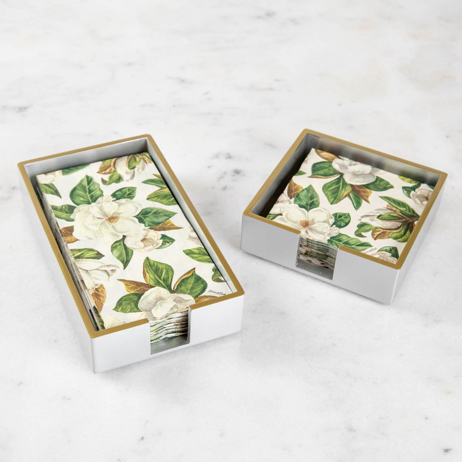 A silver colored lacquered napkin holder in guest and cocktail size with napkins in them. 