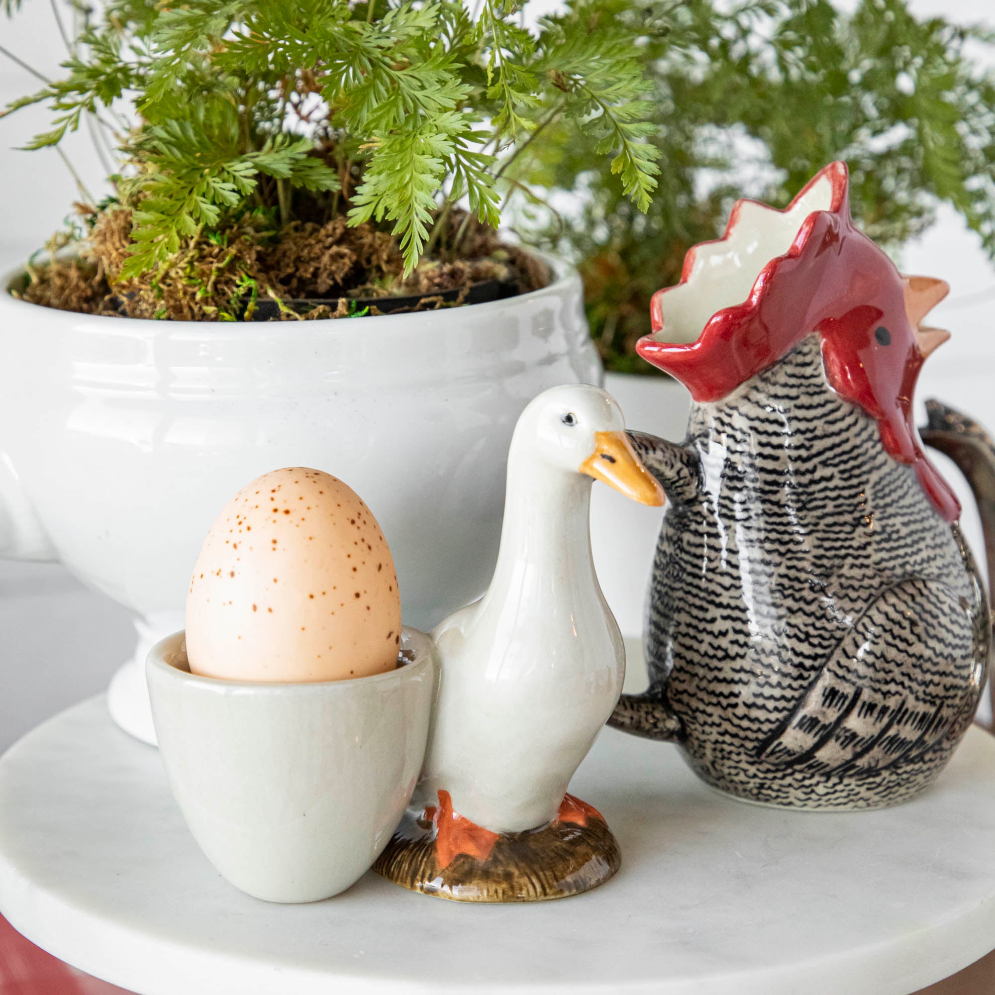 A quirky Farm Animal Ceramic rooster by British brand, Quail, sits next to a plant on a table.