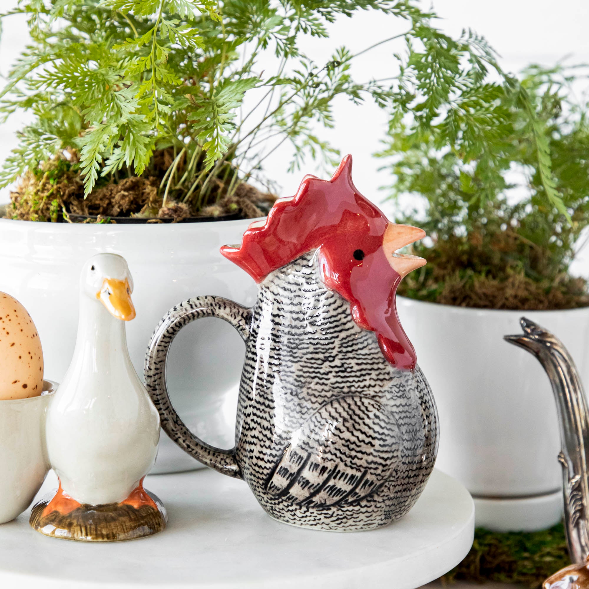 A quirky Farm Animal Ceramics mug from Quail, along with a plate of delicious eggs and a vibrant potted plant, all displayed on a table.