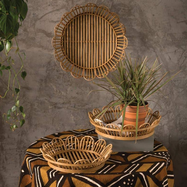 A collection of Raffi Natural Fiber Trays and a potted plant arranged on a table with a patterned cloth, against a textured grey wall with hanging greenery. (Brand: Accent Decor)