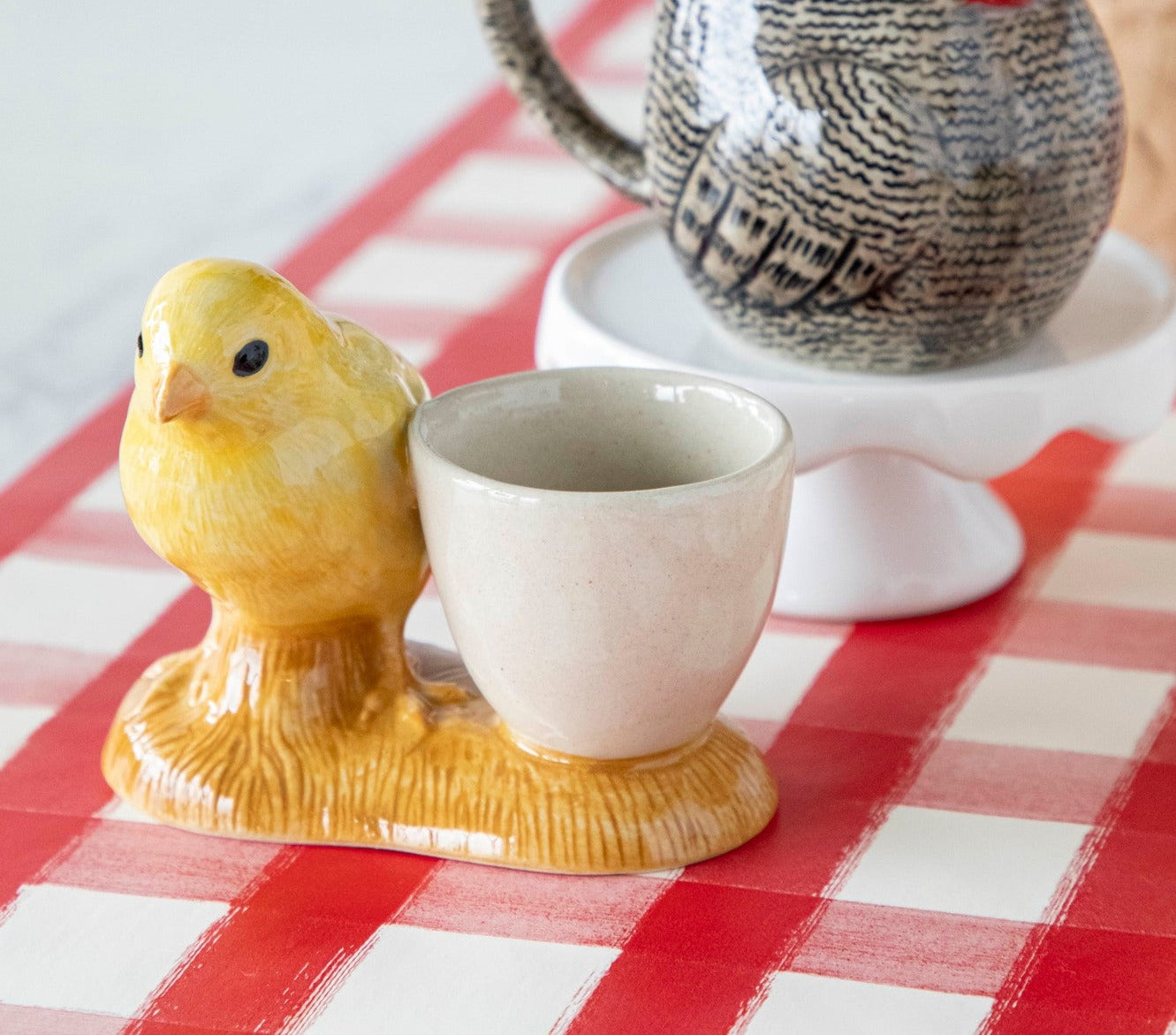 A yellow chicken, one of the Farm Animal Ceramics from Quail, sits on a red and white checkered tablecloth.
