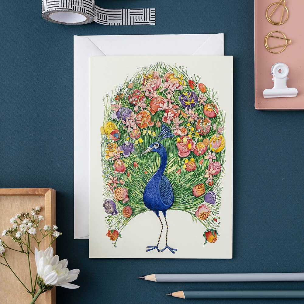Illustrated watercolour painting The DM Collection peacock greeting card surrounded by artistic supplies on a desk.