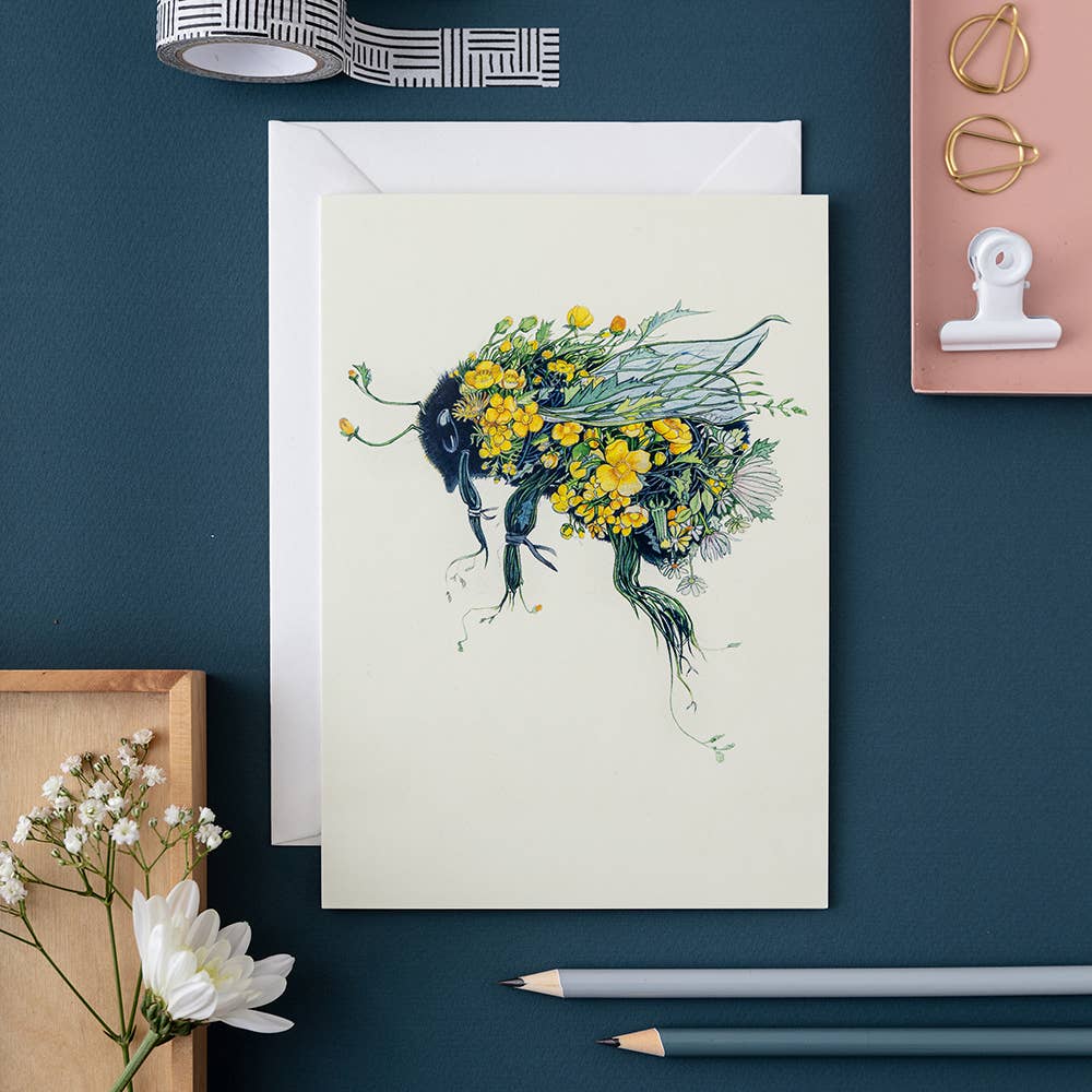 Bumblebee Card from The DM Collection with a watercolour floral bee illustration on a workspace accompanied by stationery items.