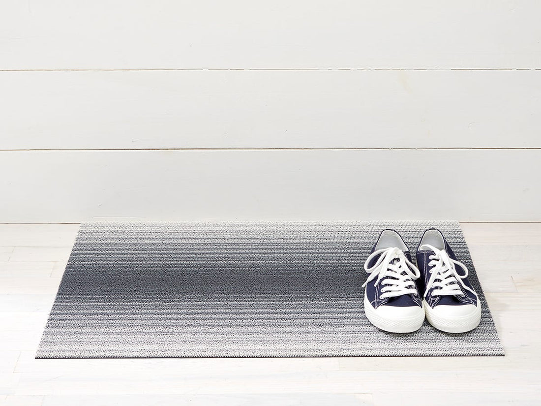 A pair of Fade Stripe Shag Utility Mat, Stone sneakers and a Chilewich door mat with ombré effect on a white floor.