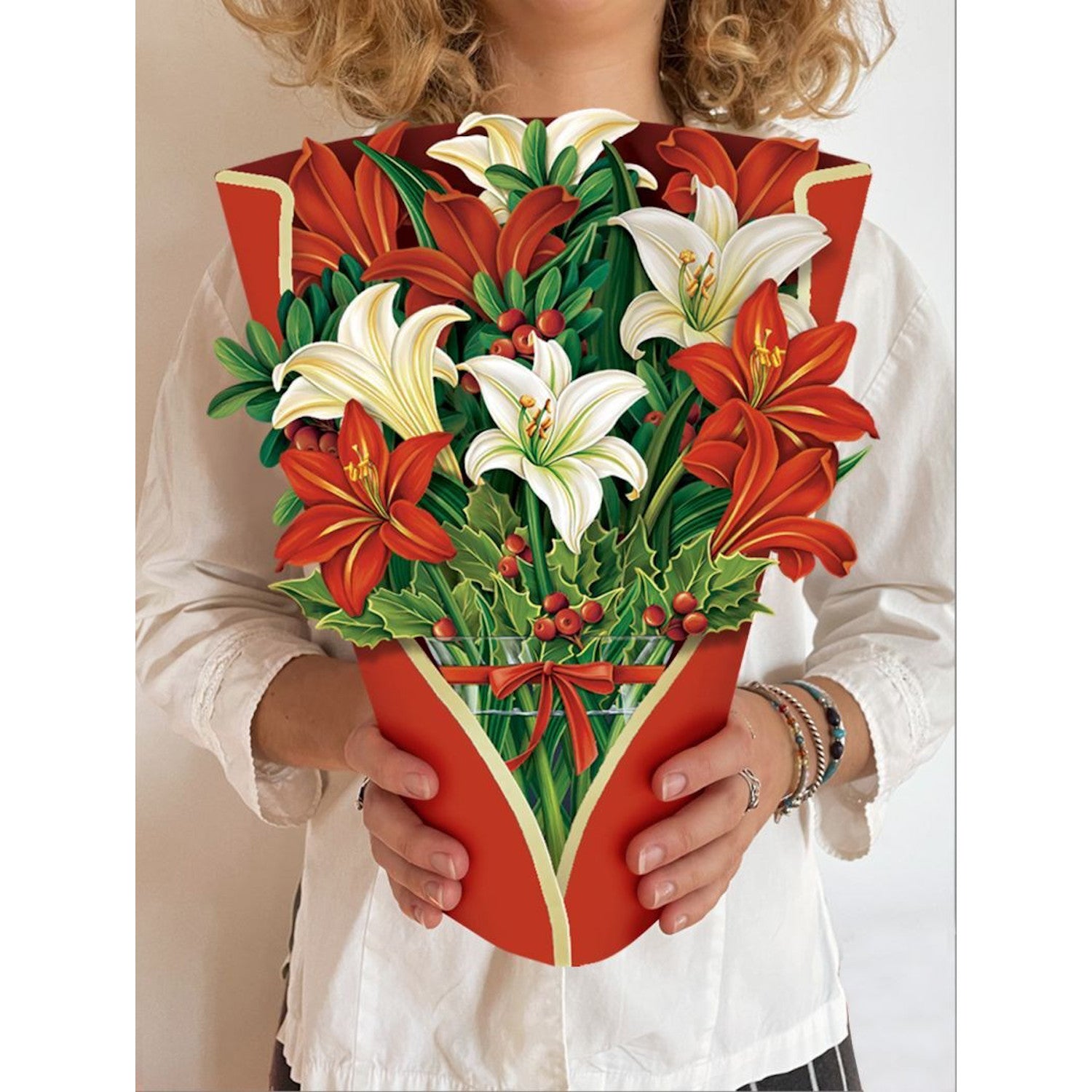 A person holding a large Winter Joy Pop Up Flower Bouquet of white lilies, Amaryllis, and holly branches wrapped in red paper from Fresh Cut Paper.