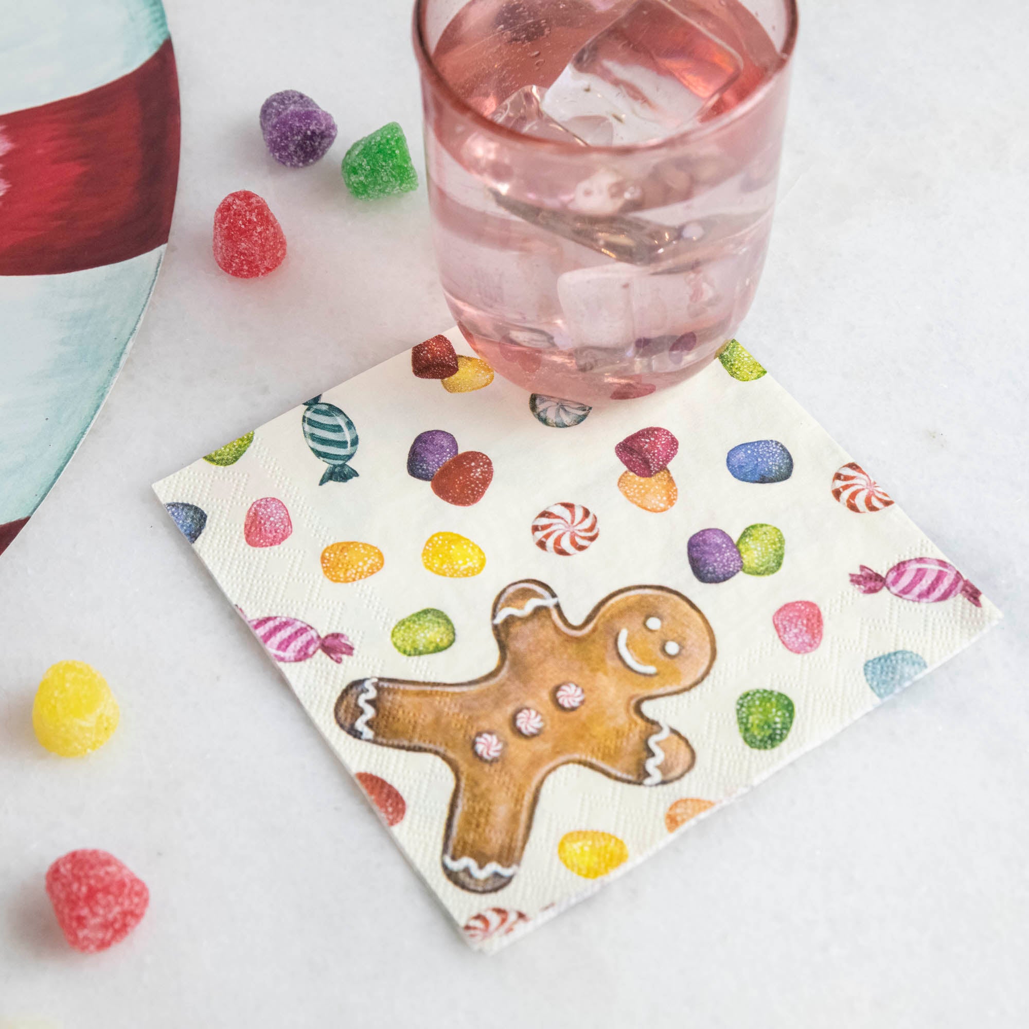 A Gingerbread Cocktail Napkin under a pink water glass, on a Christmas candy-themed table.