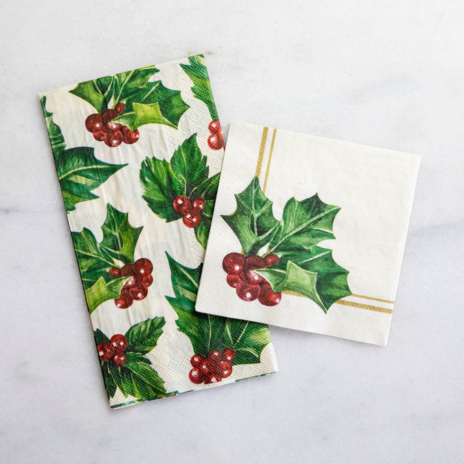 A square cocktail napkin featuring painterly green holly leaves and red berries in the lower right, with decorative gold frame lines along the lower and right edges, on a white background.