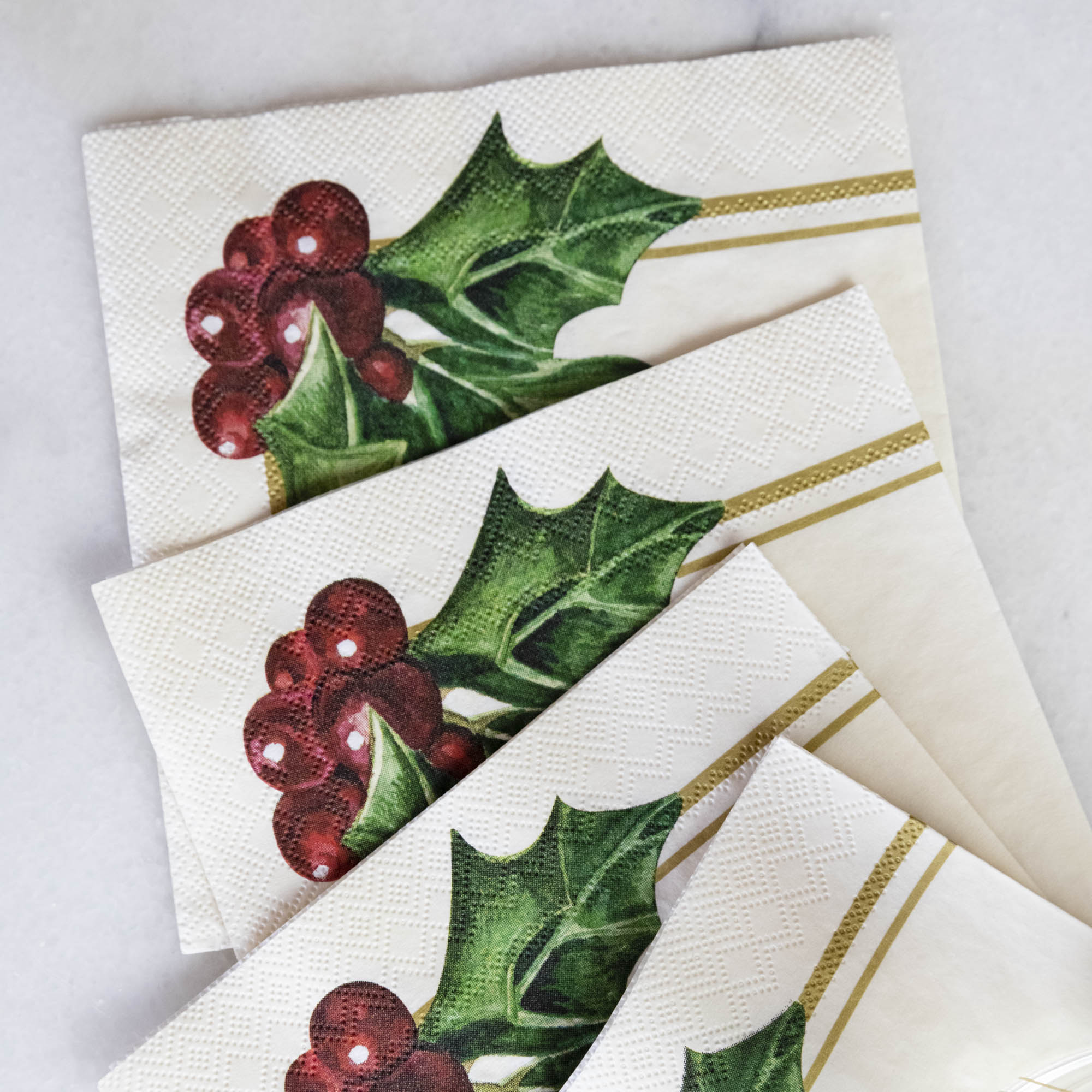 A square cocktail napkin featuring painterly green holly leaves and red berries in the lower right, with decorative gold frame lines along the lower and right edges, on a white background.