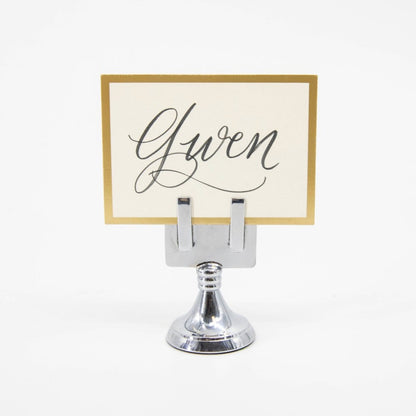 An elegant, chrome, short round stand topped by a flat card-holder with two flat prongs, holding a place card reading &quot;Gwen&quot;.
