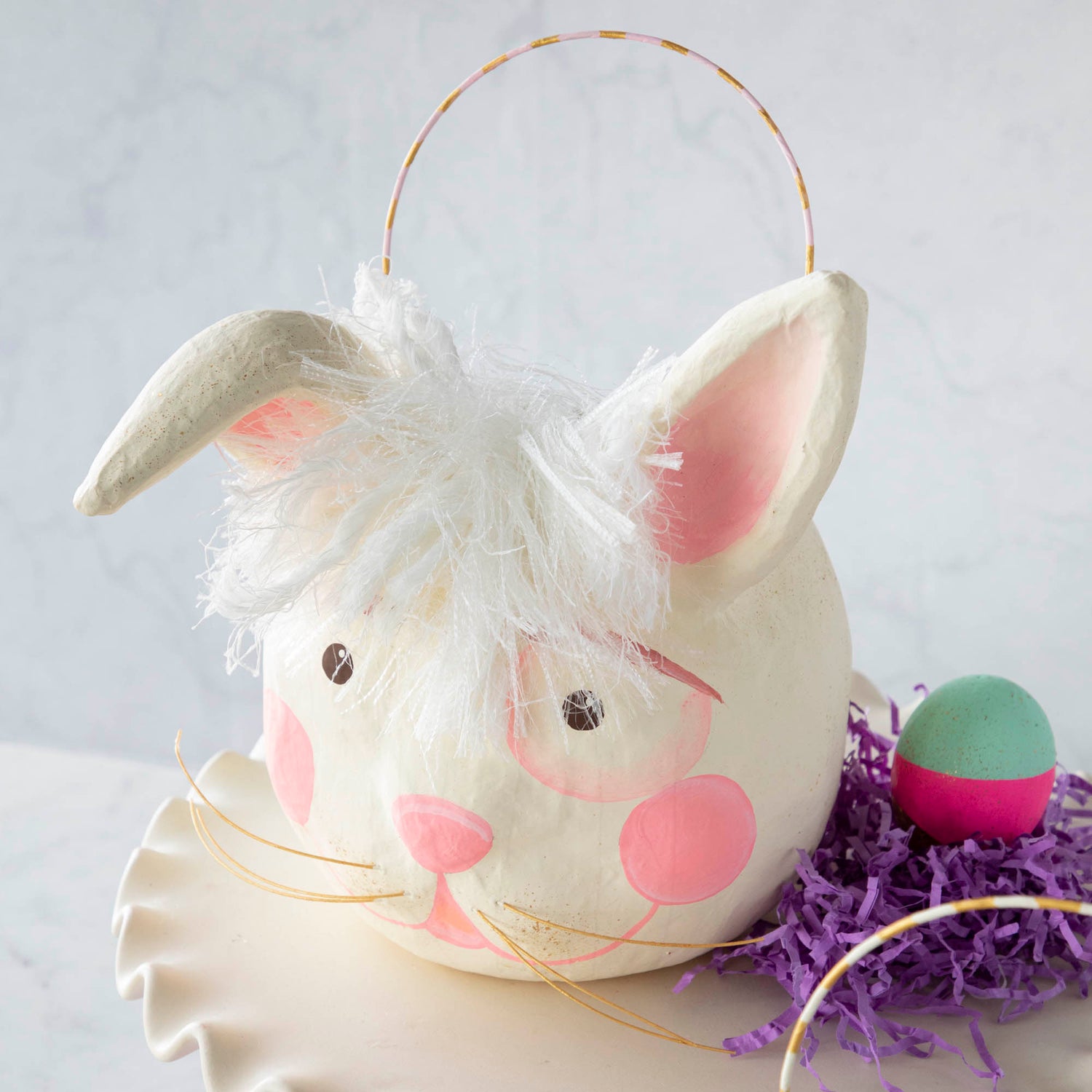 A Glitterville Papermache Easter Bucket on a cake stand, perfect for Easter fun and spring decor.